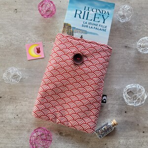 Könnte beinhalten: Roter Buchumschlag mit einem weißen Wellenmuster und einem Knopfverschluss. Der Buchumschlag ist aus Stoff und hat ein schwarzes Etikett mit einem weißen Buchstaben C. Der Buchtitel lautet "Lucinda Riley" und "La Jeune Fille Sur La Falaise".
