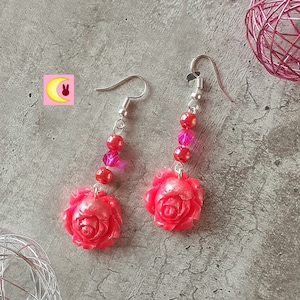 Puede incluir: Un par de pendientes colgantes con un diseño de rosa roja. Los pendientes tienen ganchos plateados y están adornados con cuentas rojas y rosas. Las rosas tienen una apariencia texturizada.