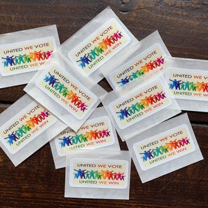 May include: A collection of rectangular stickers in small, translucent packets. Each sticker features the text "UNITED WE VOTE UNITED WE WIN" above a graphic of stick figures in a rainbow of colors, all holding hands. The stickers are on a dark wood surface.