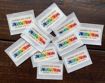 May include: Several small packets containing rectangular stickers. Each sticker displays the text "UNITED WE VOTE UNITED WE WIN" above a graphic of colourful stick figures holding hands. The stickers are arranged on a dark wooden surface.