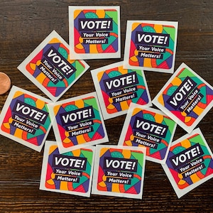 May include: A collection of square stickers with the words "VOTE! Your Voice Matters!" in a bold, colorful design. The stickers feature a vibrant palette of purple, green, orange, and red, with a white background. A penny is visible in the image.