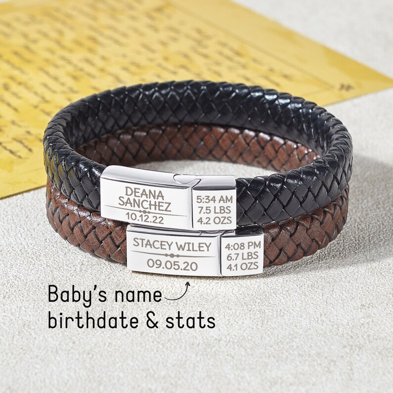 Puede incluir: Dos pulseras de cuero trenzado, una negra y otra marr&oacute;n, con cierres plateados. Los cierres est&aacute;n grabados con nombres, fechas de nacimiento y estad&iacute;sticas del beb&eacute;, incluido el peso en libras y onzas. Tambi&eacute;n se ve el texto "Baby's name birthdate & stats".
