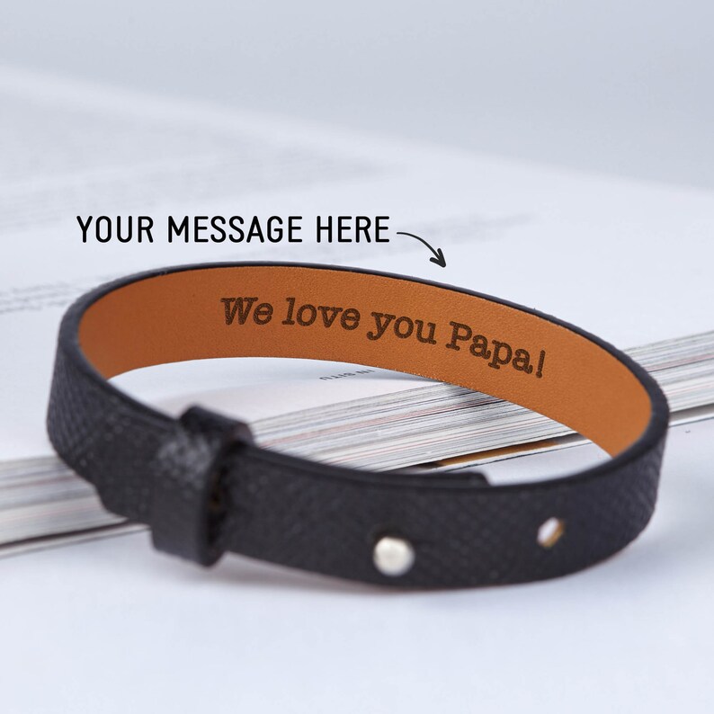 May include: Black leather bracelet with a brown leather interior. The bracelet has a silver rivet and is engraved with the text "We love you Papa!"