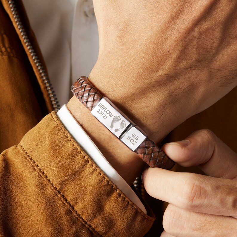 Puede incluir: Pulsera trenzada de cuero marr&oacute;n con una placa plateada grabada con "HARLOW 2.10.22" y una huella de pie, y otra placa plateada grabada con "6LB 15OZ".