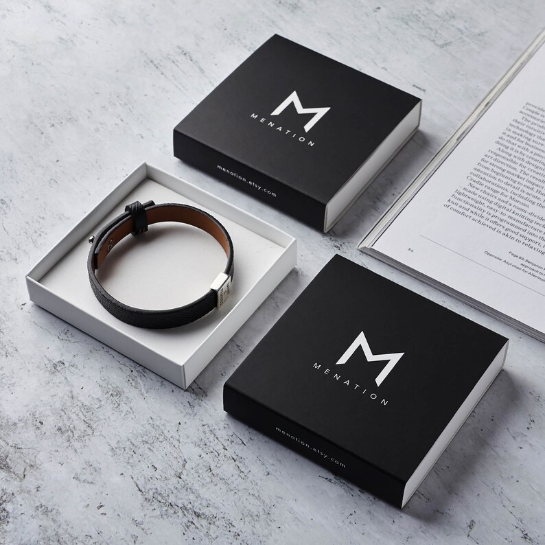 Puede incluir: Dos cajas cuadradas negras con la palabra "MENATION" y la letra "M" en blanco. Las cajas est&aacute;n sobre una superficie gris. Una caja contiene una pulsera de cuero negro con una hebilla plateada.