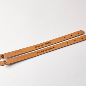 May include: Two brown leather bracelets with silver clasps. The bracelets are engraved with the text "I love you, Daddy" and "You are my hero".