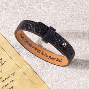 May include: Black leather bracelet with a brown leather interior. The bracelet has a silver rivet and the words "Dad, I'm so proud to be your kid." engraved on the inside.