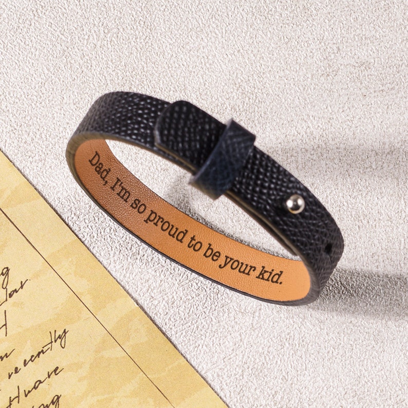 May include: Black leather bracelet with a brown leather interior. The bracelet has a silver rivet and the words "Dad, I'm so proud to be your kid." engraved on the inside.