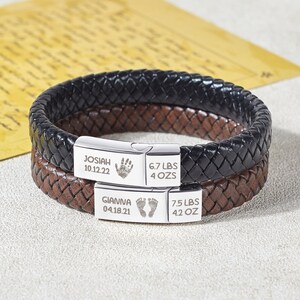 Puede incluir: Dos pulseras de cuero con placas de plata grabadas. La placa superior dice "Josiah 10.12.22 6.7 LBS 4 OZ" con una huella de mano. La placa inferior dice "Gianna 04.18.21 7.5 LBS 4.2 OZ" con huellas de pies.