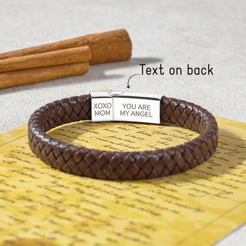 Puede incluir: Una pulsera trenzada de cuero marr&oacute;n con un cierre plateado. El cierre est&aacute; grabado con "XOXO MOM" y "YOU ARE MY ANGEL". La pulsera se muestra sobre un papel amarillento con palitos de canela en el fondo.