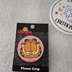 Könnte beinhalten: Ein pinkfarbener Handyhalter mit Garfield's Gesicht, orangefarbenem Fell und einem mürrischen Gesichtsausdruck. Der Handyhalter befindet sich auf einer schwarzen Rückseite mit dem Schriftzug "Phone Grip" unten. Im Hintergrund befindet sich ein weißer Aufkleber mit einem Bären und Sternen.