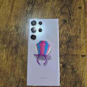 May include: A light purple smartphone with a multi-lens camera system and a pop-up phone grip. The phone grip features a pink, blue, and white geometric design with a purple ring.