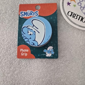 Könnte beinhalten: Ein runder Handygriff mit einer blauen Schlumpf-Figur auf hellblauem Hintergrund. Die Verpackung ist blaugrün mit orangefarbenen Akzenten und zeigt den Text The Smurfs und Phone Grip sowie eine kleine Abbildung eines rennenden Schlumpfes.