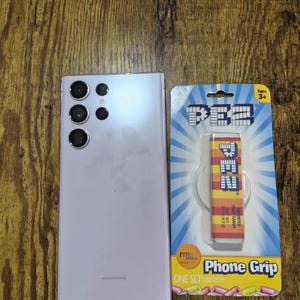 May include: A light purple smartphone with multiple rear cameras is next to a PEZ-themed phone grip. The phone grip is on a blue and yellow card that reads "Phone Grip" and "Ages 3+". The PEZ dispenser is red, orange, and yellow.