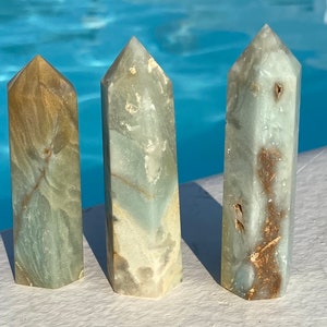 May include: Three green and brown crystal points, also known as green calcite, are arranged in a row on a white surface. The points are all different sizes and shapes, but they are all polished to a smooth finish.