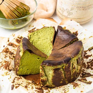 May include: A sliced matcha cheesecake with a dark, caramelized crust. The cake sits on parchment paper, with a bowl of matcha and a whisk visible in the background. The cake has a vibrant green interior.