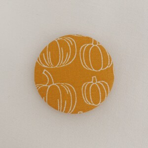 May include: A round, fabric button with a mustard yellow background and a white pumpkin pattern. The button is a seasonal item, perfect for fall-themed crafts and decorations.