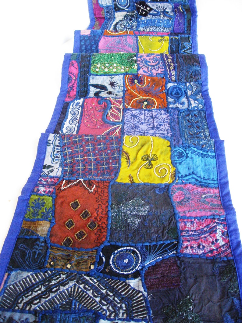 Peut inclure: Un long chemin de table rectangulaire en patchwork avec une bordure bleue. Le chemin de table est compos&eacute; de diff&eacute;rents tissus de couleurs et de motifs vari&eacute;s, notamment rouge, vert, jaune et violet. Le chemin de table est d&eacute;cor&eacute; de perles et de paillettes.