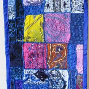 Peut inclure: Une courtepointe patchwork avec une bordure bleue et des carr&eacute;s de tissu de diff&eacute;rentes couleurs. La courtepointe pr&eacute;sente des broderies et des perles complexes dans des tons de bleu, rose, jaune et marron.