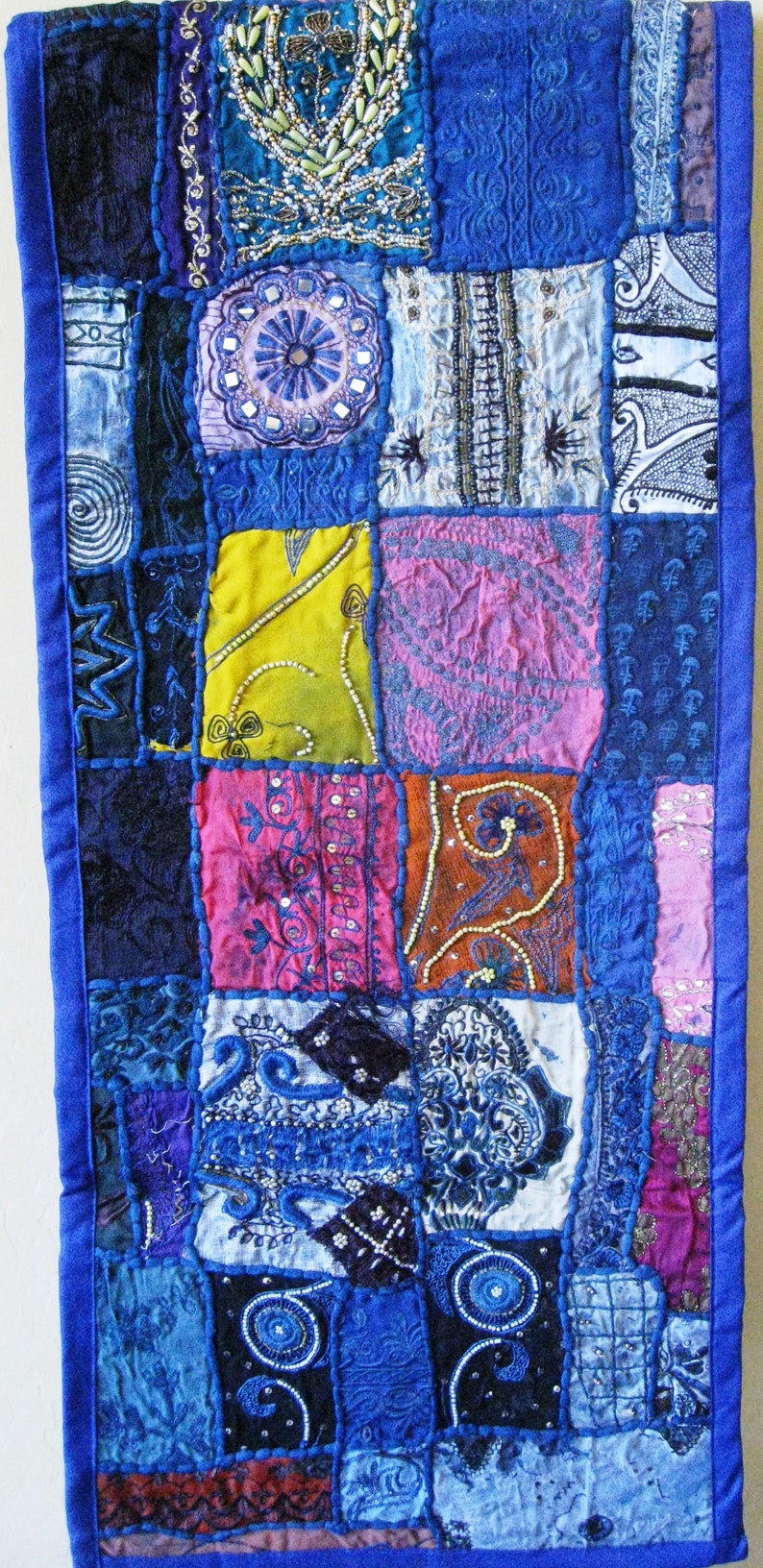 Peut inclure: Une courtepointe patchwork avec une bordure bleue et des carr&eacute;s de tissu de diff&eacute;rentes couleurs. La courtepointe pr&eacute;sente des broderies et des perles complexes dans des tons de bleu, rose, jaune et marron.