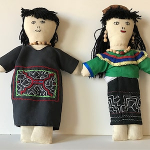 May include: Two handmade fabric dolls wearing traditional clothing. The doll on the left is wearing a black dress with red and green embroidery. The doll on the right is wearing a green shirt with blue and yellow trim and a black skirt with white embroidery.
