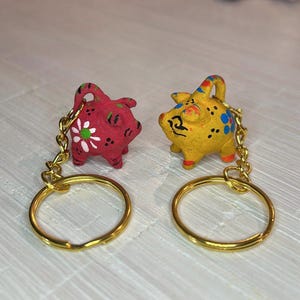 Peut inclure: Deux petits porte-clés en forme de cochon peints à la main. L'un est rouge avec une fleur blanche, l'autre jaune avec des pois bleus. Les deux ont des anneaux et des chaînes dorés. Ils sont probablement en céramique.