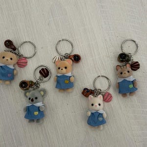 Peut inclure: Une collection de six porte-clés sur le thème des animaux. Chaque porte-clés présente une petite figurine d'animal en peluche, dont un ours, un koala et un chat, vêtue d'une robe bleue. Les porte-clés ont des anneaux argentés et des breloques décoratives.