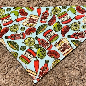 May include: A blue bandana with a white background and a pattern of red, green, and yellow chili peppers, limes, avocados, and jars of salsa and hot sauce. The text on the jars reads "Salsa", "Hot", "Chili", "Queso", and "Nacho Chips".
