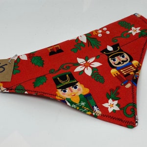 May include: Red fabric dog bandana with a Christmas nutcracker pattern. The bandana has a black stitched edge and features white flowers, green holly leaves, and red berries. The text "XS" is written on a brown tag.