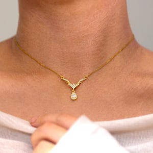 May include: Gold necklace with a delicate design. The necklace features a V-shaped pendant adorned with small, sparkling stones and a teardrop-shaped charm. The jewelry is worn on a person's neck, against a white top.