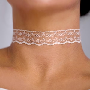 Puede incluir: Una gargantilla de encaje blanco con un delicado patrón floral y un borde festoneado. El accesorio se ajusta al cuello, mostrando un intrincado trabajo de aguja y una estructura ligera y semitransparente en toda la banda.