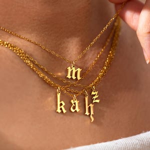 May include: Gold-colored layered necklaces with initial pendants. The necklaces are worn around the neck, with the initials "m", "k", "a", "h", and "z" hanging from the chains. The chains are delicate and the pendants are in a gothic font.