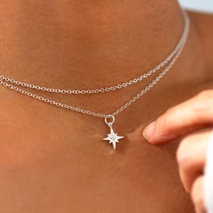May include: A silver-colored, layered necklace with a starburst pendant. The necklace is composed of two delicate chains, with a small star-shaped charm at the center. The starburst pendant has a central stone and is worn on the neck.
