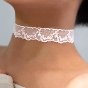 May include: A delicate, light pink lace choker necklace. The choker features a scalloped edge and floral detailing. The necklace is worn around the neck, showcasing its intricate design and soft color. A stylish accessory for any occasion.