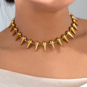 May include: A gold-colored choker necklace featuring a series of pointed, cone-shaped pendants. The necklace is made of small, linked chain segments. The necklace is worn around the neck.