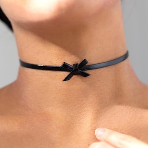 May include: A black choker necklace with a small bow detail. The choker is made of a thin, shiny material and sits close to the neck. The bow is centered at the front of the choker.
