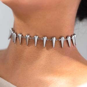 May include: Silver-toned choker necklace featuring a series of cone-shaped spikes. The necklace is worn around the neck, with the spikes evenly spaced along a delicate chain. The jewelry is a bold statement piece.