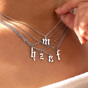 May include: A silver-colored layered necklace with multiple chains. Each chain has a different pendant with a letter in an old English font. The letters are 'm', 'h', 'z', 'b', 'f'. The necklace is worn around the neck.