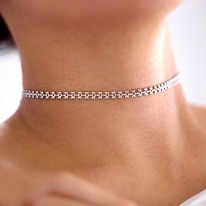 May include: A silver choker necklace with a delicate, repeating star-like pattern. The necklace sits around the neck, showcasing a minimalist and elegant design. The jewelry is a stylish accessory.