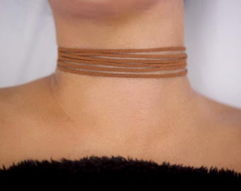 May include: A brown suede choker necklace with multiple strands. The necklace is wrapped around the neck, creating a layered effect. The choker is a neutral color and can be paired with various outfits.