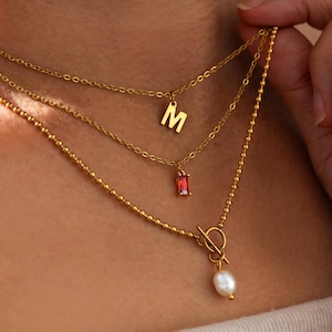 May include: Gold-coloured layered necklaces. The top chain has a letter "M" pendant, the middle chain has a red rectangular gem, and the bottom chain has a pearl pendant. The necklaces are worn around the neck.