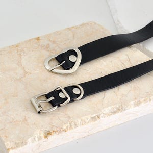 Pu&ograve; includere: Due collane choker in pelle nera con fibbie argentate a forma di cuore e quadrate.