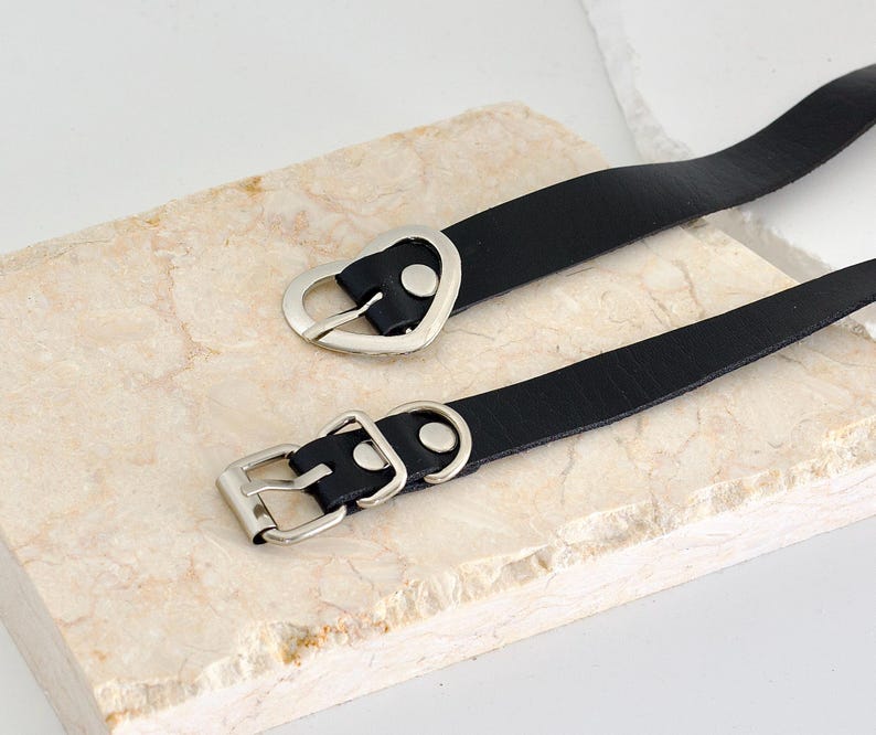 Pu&ograve; includere: Due collane choker in pelle nera con fibbie argentate a forma di cuore e quadrate.