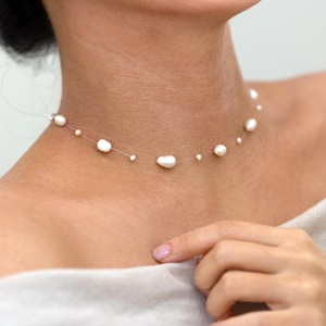 May include: A delicate pearl necklace, with irregularly shaped white pearls strung on a fine, almost invisible thread. The necklace is shown against a neutral background, emphasising the elegant simplicity of the jewellery.