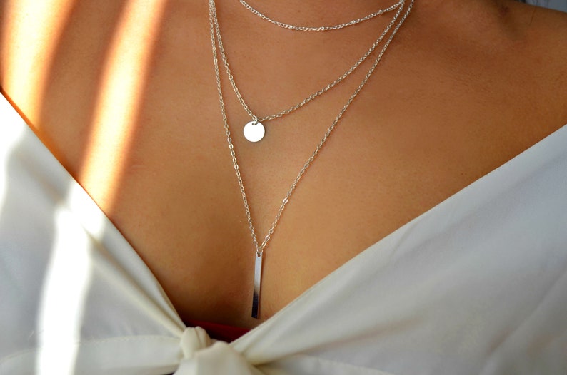 May include: A silver layered necklace with a round pendant and a long, thin bar pendant.