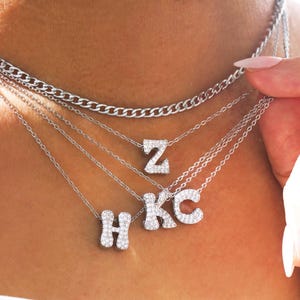 May include: A collection of silver necklaces with initial pendants. The necklaces feature a variety of chain styles, including a curb chain and delicate link chains. The initial pendants are encrusted with small, sparkling stones.