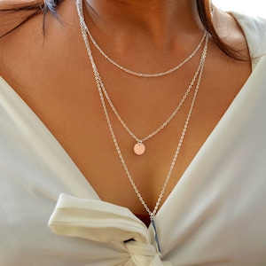 May include: A silver layered necklace with a round pendant. The necklace is worn by a person with a white shirt.