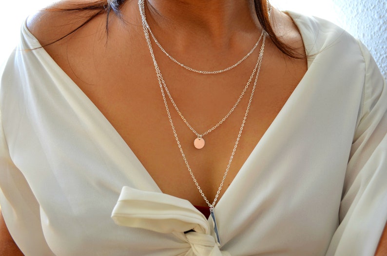 May include: A silver layered necklace with a round pendant. The necklace is worn by a person with a white shirt.