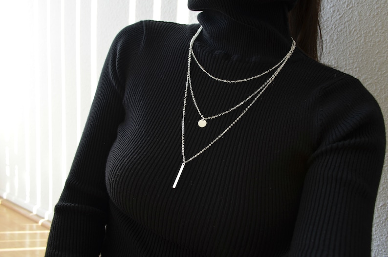 May include: A layered silver necklace with a small round pendant and a long, thin bar pendant.