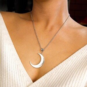May include: A silver crescent moon pendant necklace on a silver chain. The necklace is worn by a person with a white sweater.